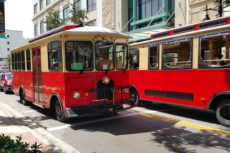 Two red trolleys pass each other on the streets of West Palm Beach. One of the trolleys has a sign in the window that reads “green line.” 