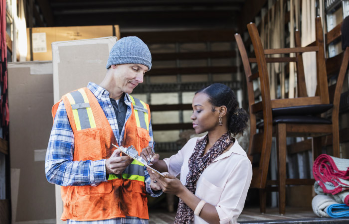 A woman is standing behind a partially loaded moving truck, signing moving insurance paperwork while the mover holds the clipboard.