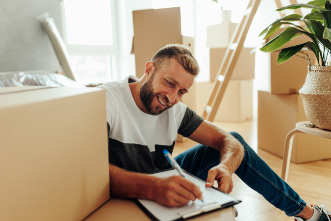 A happy man is sitting on the floor surrounded by moving boxes and furniture as he checks items off of his moving checklist.