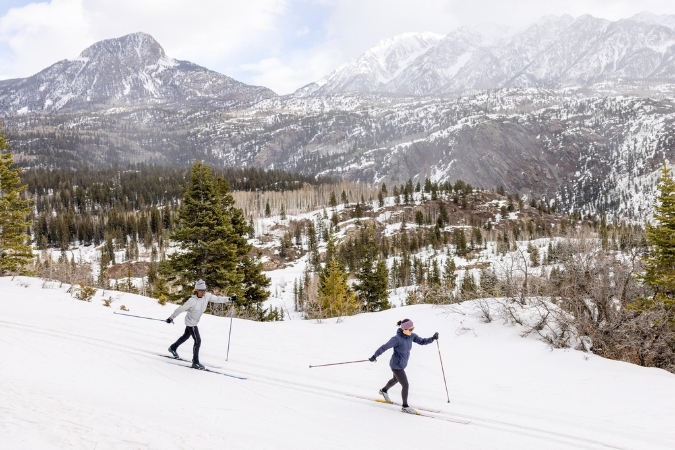 Two skiers living in Durango, CO, glide down a mountainside surrounded by snowcapped peaks and scattered evergreen firs.