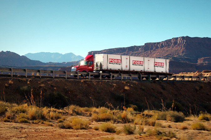 A PODS truck is transporting three PODS portable moving and storage containers through an arid landscape in the western United States. 