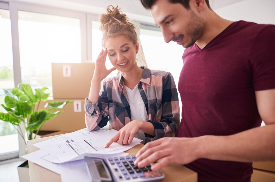 A couple is moving on a budget, so they’re working out the numbers using a calculator in their living room surrounded by moving boxes.