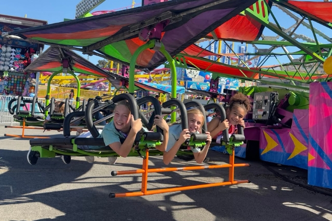 Children living in Little Rock, Arkansas, are having fun riding a colorful carnival attraction at the Arkansas State Fair with games and more carnival stalls visible in the background.