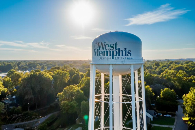 A sunny day view of a water tower in West Memphis, Arkansas, surrounded by lush greenery and blue skies.