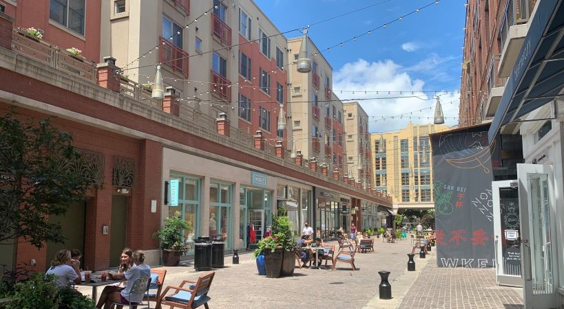 View of a downtown street in Bethesda, Maryland — a popular DC suburb. Locals are out and about, enjoying a sunny day in the city. Some are dining al fresco while others are strolling by storefronts.