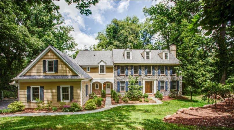 Front view of a yellow siding and stone, two-story home in Newtown Square, Pennsylvania — a city close to Philadelphia — with a well-manicured lawn and mature trees casting shadows on the house. 