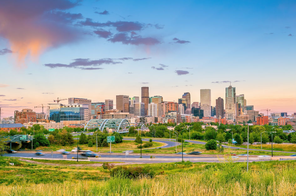 The skyline of Denver, Colorado. There are skyscrapers and other buildings. There are a few wispy clouds in the sky and green grass in the foreground.