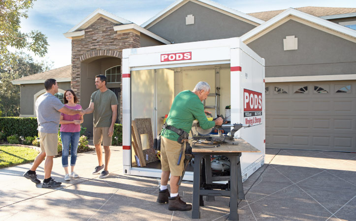 A couple is speaking with a contractor in their driveway. A second contractor is working near a PODS container that is being used to store tools and materials during the primary bathroom remodel.