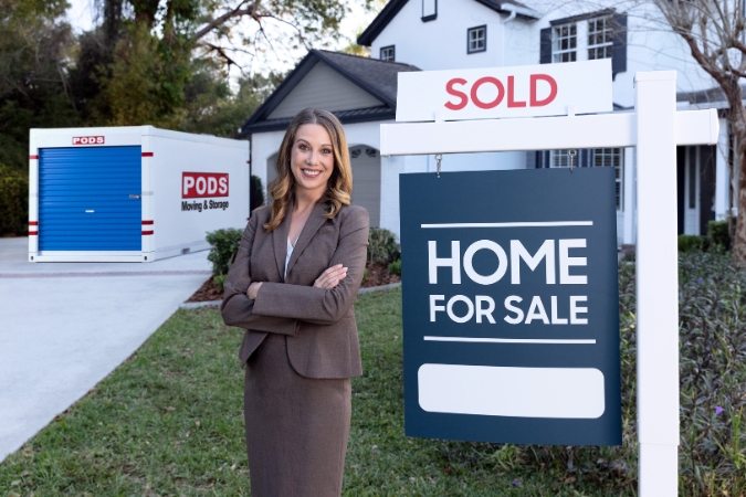 A real estate agent is standing next to a “For Sale” sign outside a new home with a PODS portable container in the driveway.