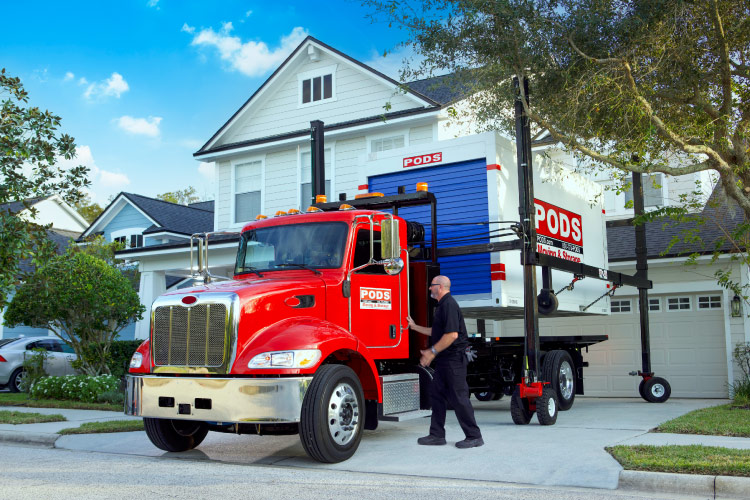 A PODS driver is about to climb back into his truck to move it out from under PODZILLA, which is holding a PODS container aloft. After he moves the truck, he’ll use PODZILLA to safely place the PODS container in the driveway, so it can easily be loaded.