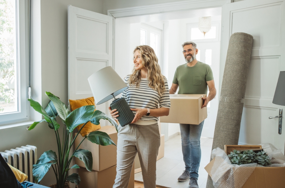 A mature couple is carrying things into the entrance of their new house, which is littered with other items, including a rolled-up rug, house plants, and moving boxes.