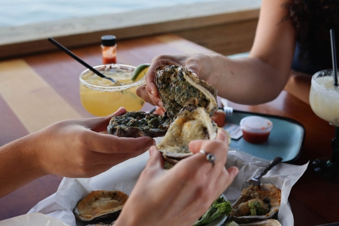 Three people cheer with grilled oysters over a table at an oyster bar in North Port, FL, with cocktails in the background.