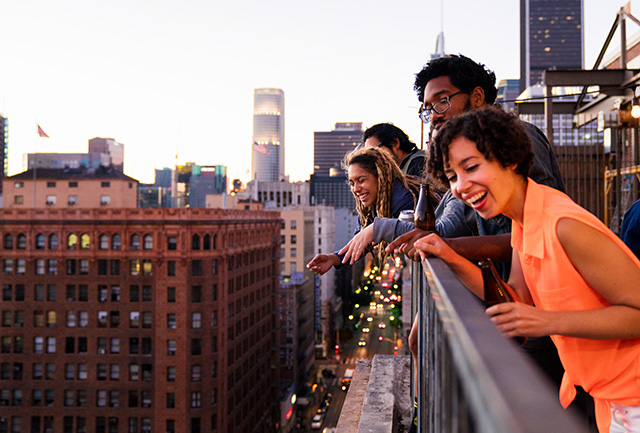 A group of four friends stand on a balcony and look out into the city of Los Angeles. Below them is traffic and in the background there is a brown building.