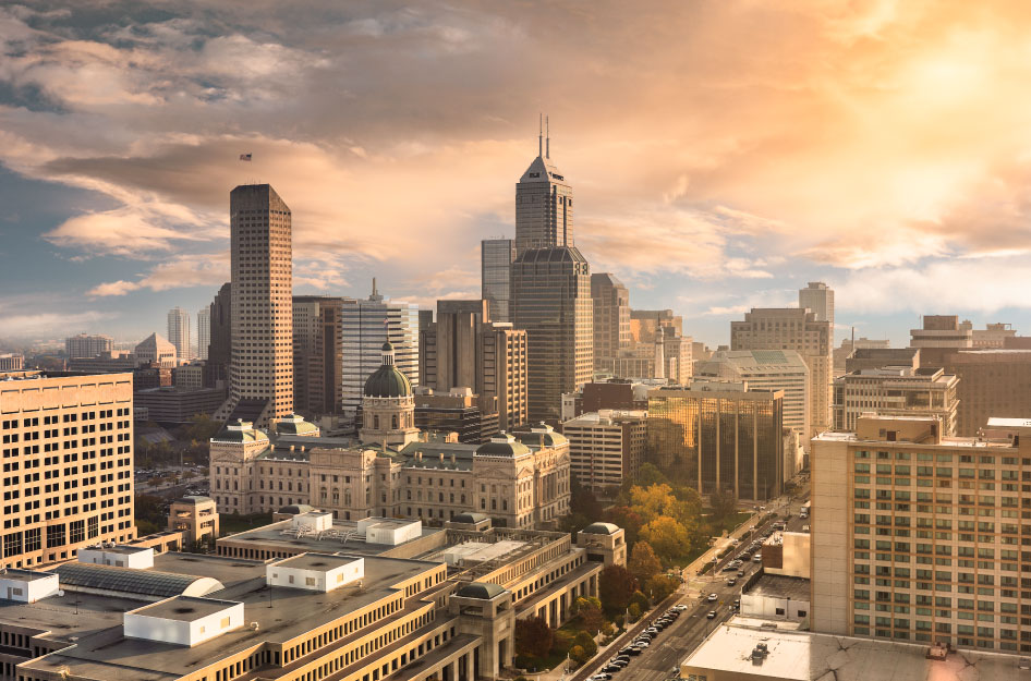 Rays of light are shining through a rainbow of clouds onto the buildings of downtown Indianapolis during sunset.