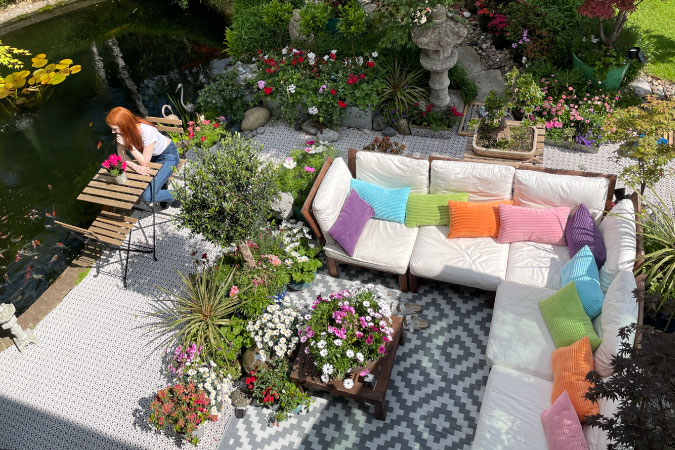 A woman is relaxing in a cozy sitting area beside a koi pond in her backyard surrounded by lush landscaping.