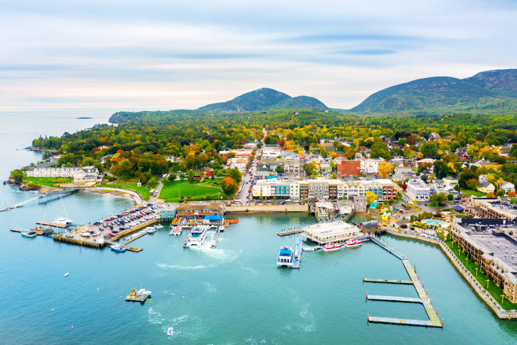 Aerial view of Bar Harbor in Maine. There is a large marina along the coast. The city is small but dense and is surrounded by forests and low mountains.