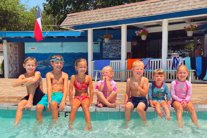 7 young kids are sitting on the edge of a community pool in Plano, Texas, on a sunny summer day. 