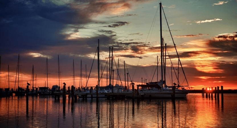 A sunset view of the Corpus Christi Marina, featuring sailboat silhouettes against a darkening sky with vibrant orange hues reflecting across the water.
