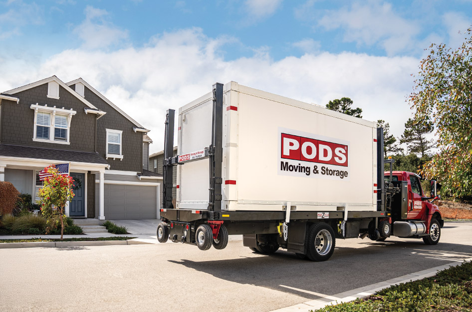 A PODS truck with a PODS portable moving container on the back is positioned outside a residential home, preparing to drop-off the container.