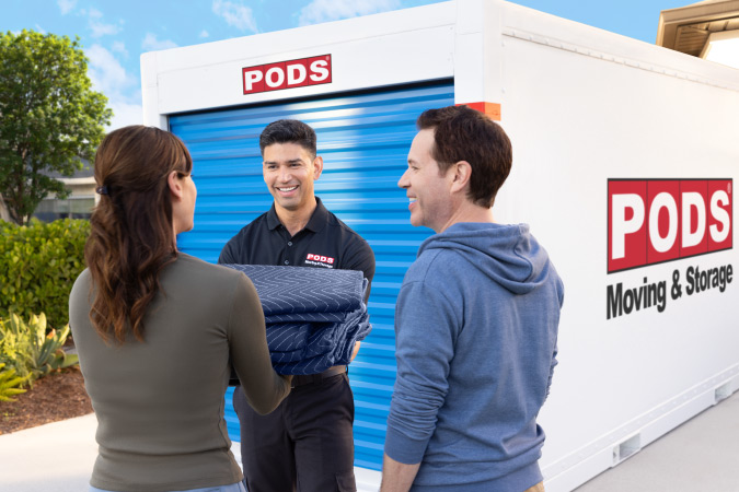 A PODS driver is handing moving blankets to a couple as they all stand in front of a PODS portable moving container in their driveway.