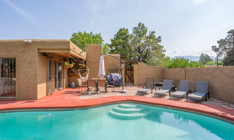 The backyard of a stucco home in the Eastside Albuquerque neighborhood of Hodgin. The patio is painted a natural terracotta color which contrasts beautifully with the blue waters of the private pool. Beyond the stucco wall surrounding the backyard, you can see lush green trees and distant mountains.
