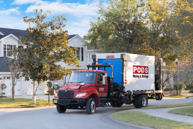 A PODS truck is transporting a portable moving and storage container through a residential neighborhood.