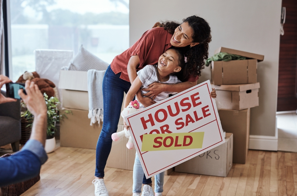 A mother and daughter are posing for a photo in front of moving boxes while holding a "Sold" real estate sign to celebrate completing all the home-buying steps.