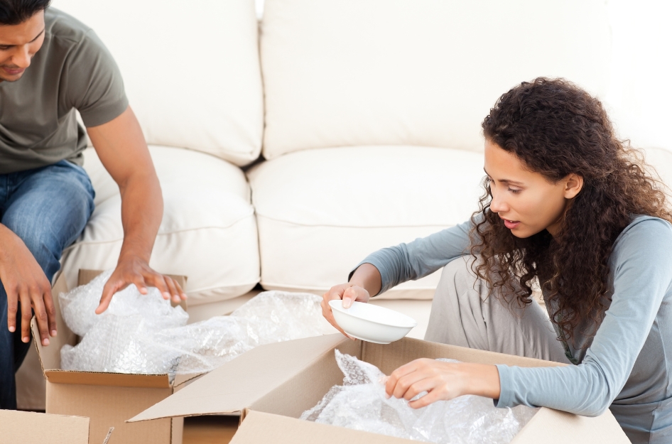 A couple is packing fragile items for moving, using bubble cushioning roll to wrap ceramic bowls before placing them into moving boxes.