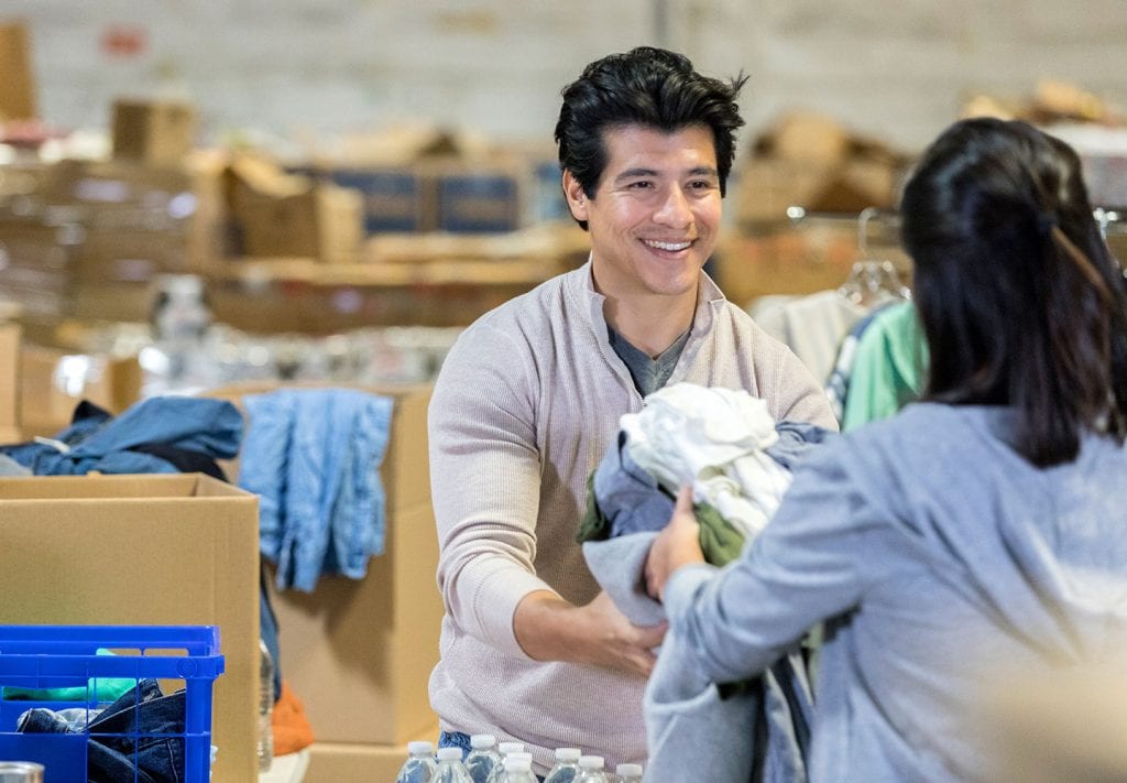 A young man accepts a dopnation of clothes from someone in a clothing distribution center. Behind him are rows of cardboard boxes.