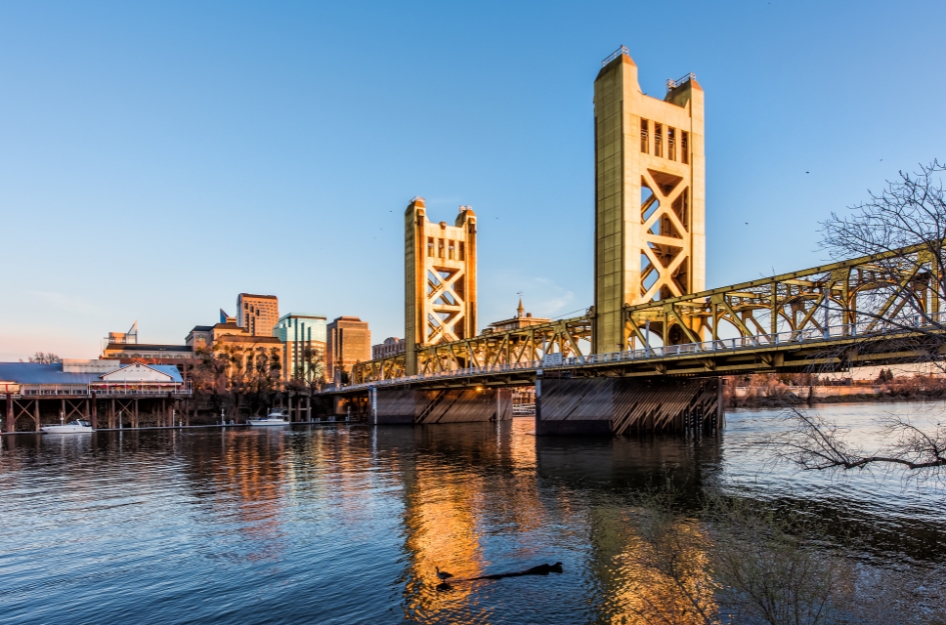 Sunrise view of Tower Bridge reflecting off of the river with the city skyline in the distance — a beautiful sight for those living in Sacramento, CA.