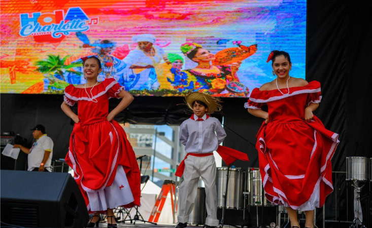 Hispanic dancers are performing on a stage in front of a screen that says “Hola Charlotte” during one of the many festivals the city offers, a major pro of living in Charlotte, NC.