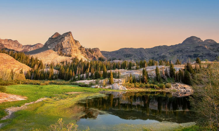 A view of the Lake Blanche Trail outside Salt Lake City, Utah, as it winds around a body of water. There are mountains and rocky formations in the distance, as well as trees sprouting up here and there.