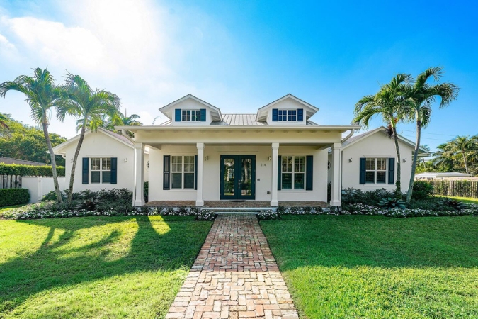 A white, one-story home in Lake Ida — one of the best places to live in Delray Beach — featuring a wide front porch, brick walkway, manicured lawn, and palm trees.