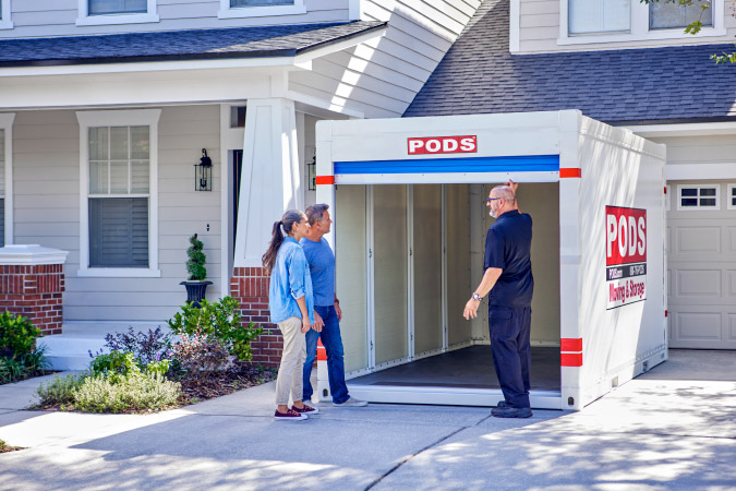 A PODS driver is opening the blue roll-up door of a PODS portable moving container to show a couple the inside of the container.