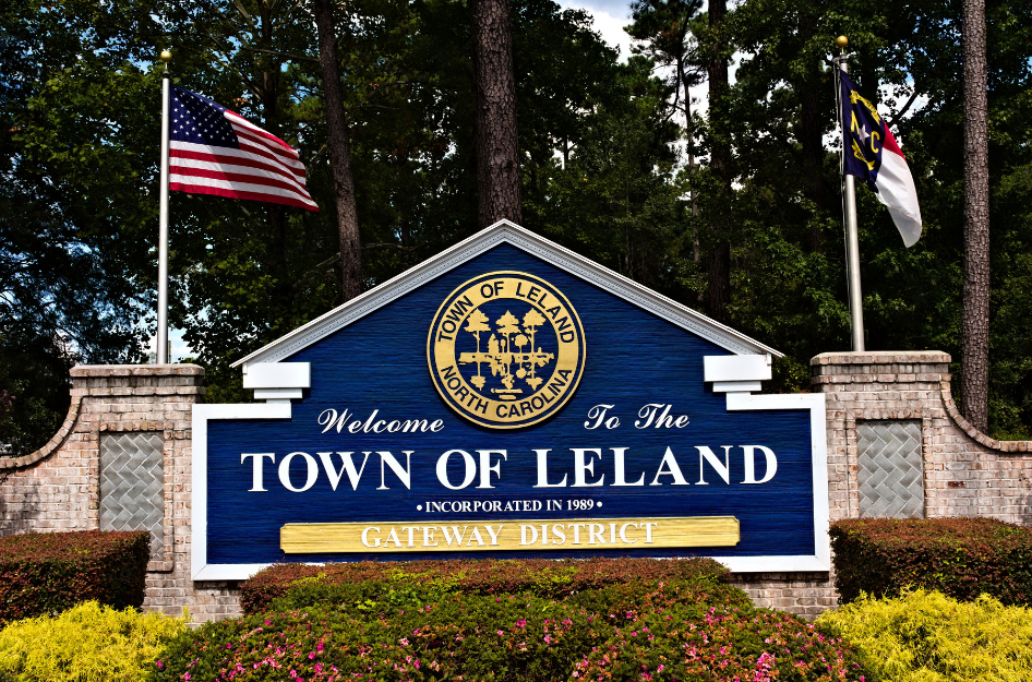 A blue wooden sign in the shape of a home that reads "Welcome to the town of Leland", a common sight when living in Leland, NC.
