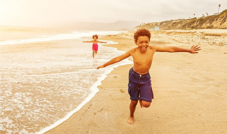 Two kids, a boy and a girl, are squealing with delight as they run down the beach with their arms outspread, pretending to fly. The water splashes around the girl’s legs as she runs.