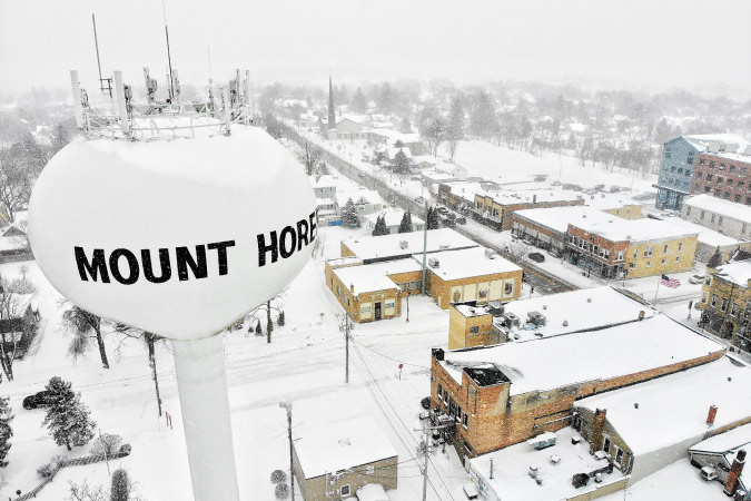 Aerial view of Mount Horeb, Wisconsin, during a blizzard, featuring short city buildings under a blanket of snow and a large water tower in the foreground.