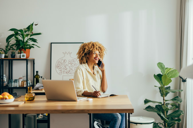 A happy woman is talking on the phone at her desk, surrounded by beautiful plants — new additions to her home office setup.