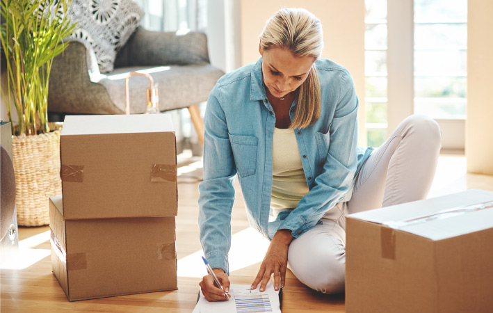 A woman sits beside some packed cardboard boxes while she makes a list of the things that she’s putting in storage.