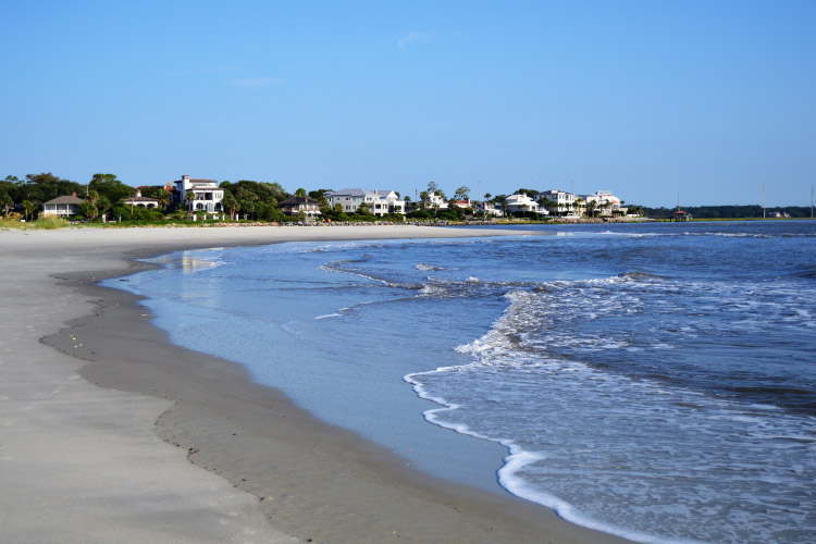 View down the beach of waterfront homes in St. Simons Island, Georgia. The sand is a light cream color, and the water is lapping gently against the shore.