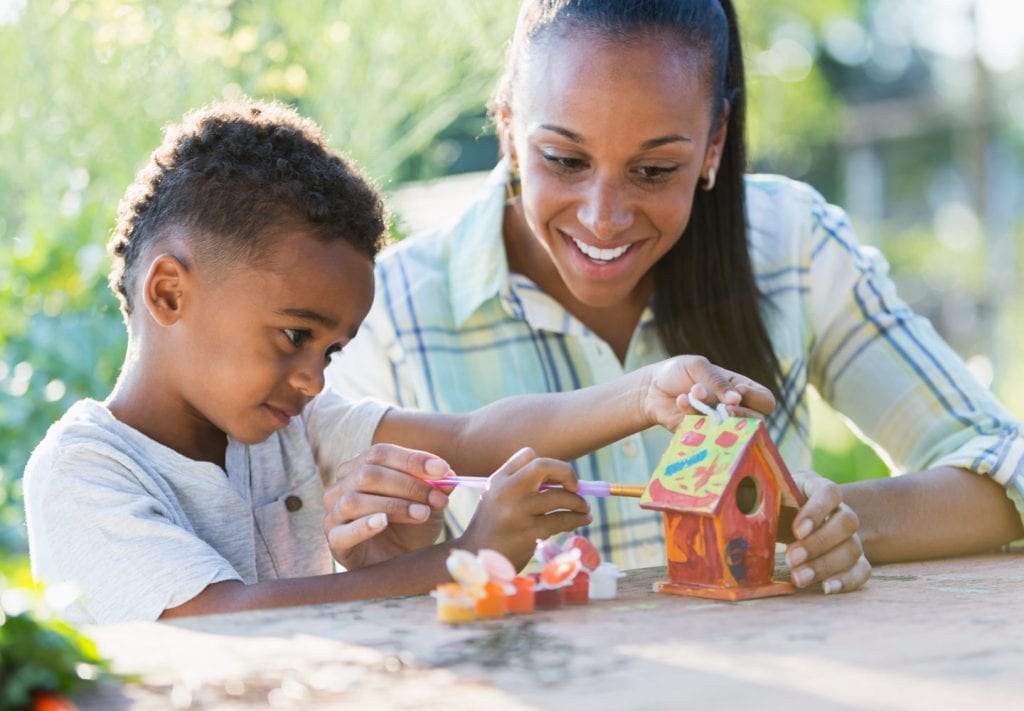 A mom and her sone are painting a bird feeder while sitting outside, enjoying the sun. 