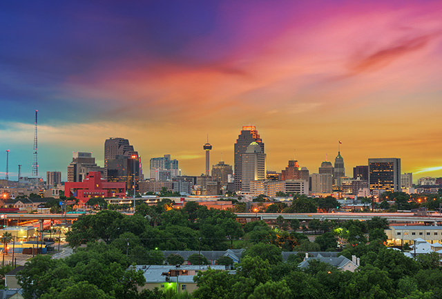 The skyline of San Antonio, Texas, is surrounded by a rainbow of clouds in the sky. There are also groups of lush, green trees in the foreground.
