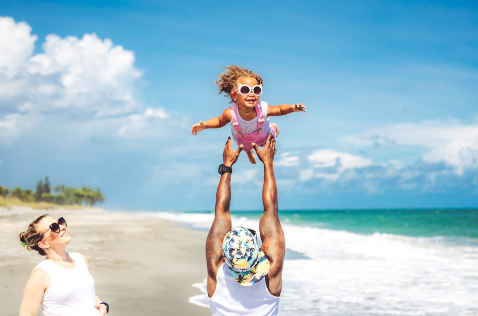 A happy girl is being lifted above her father’s head at the beach while her mother looks on — a glimpse of one of the joys of living in Florida.