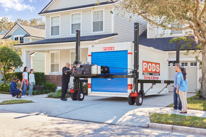 A multi-generational family is standing outside their home, watching as a PODS driver delivers their portable moving container to the driveway.