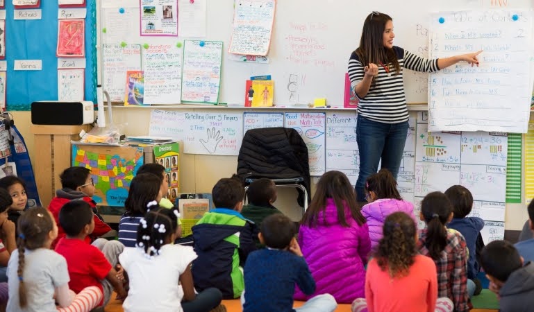 A teacher is leading her class in a lesson at a San Francisco public school. The students are sitting quietly on the floor of the classroom as the teacher directs their attention to notes on a large piece of paper hanging from the whiteboard.