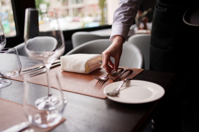 A waiter sets a dining table with polished glassware, neatly arranged place settings, and folded napkins at one of the many restaurants in Iowa.