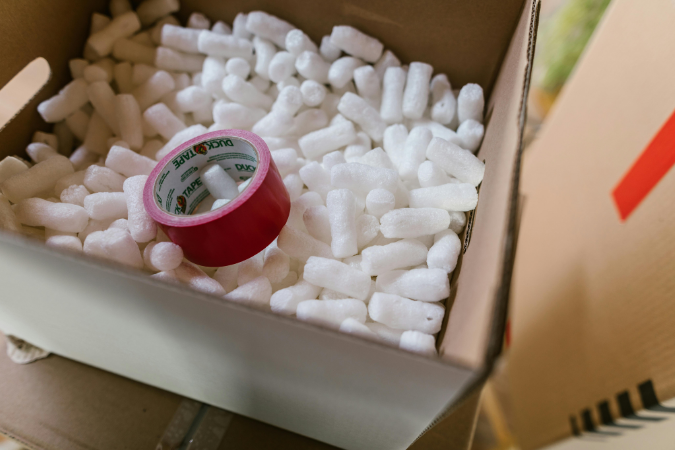 A roll of pink packing tape on top of white packing peanuts in a moving box.
