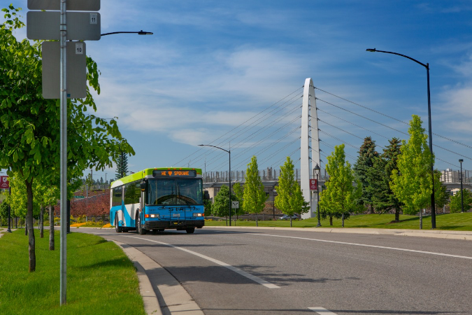 Residents living in Spokane, WA, ride a city bus down a roadway lined with trees and streetlights on a sunny day.