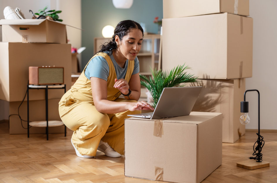 A woman is working on her laptop at a makeshift “moving box table,” surrounded by stacks of moving boxes, as she researches where to store stuff during a remote work stint.