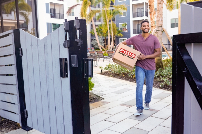 A man is smiling as he carries a moving box through the courtyard of an apartment community while moving out after a breakup.